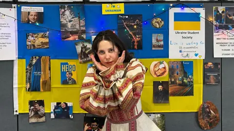 Handout A white woman is pictured in traditional Ukranian dress with her hands cupped to her face. 
She is standing in front of a Ukranian flag with pictures of Ukraine on it. 