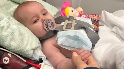 Supplied Baby Saskia is in hospital. Her mother holding her hand and there is a blanket under her head as a pillow. The baby has cooling pads on her chest and she is still strapped into an ambulance stretcher. She has a doll beside her, and a dummy in her mouth. She looks sleep, warm and unwell.