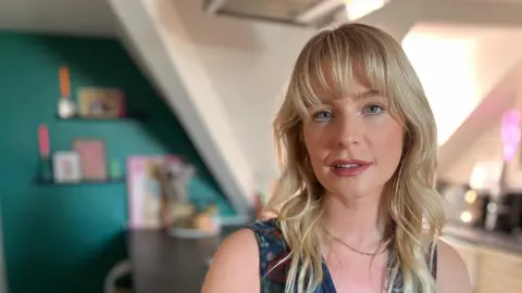 BBC Jess has blonde shoulder-length hair with a fringe and blue eyes. She is wearing
blue Aztec-design waistcoat and sitting in her kitchen.