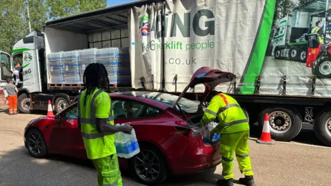 Simon Jones/BBC Two people in hi-vis jackets getting water bottles out of the back of a red car. There is a large lorry next to the car.