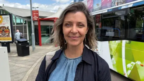 Ben Schofield/BBC Emma Pratt with mousy-brown shoulder-length hair smiles and looks at camera. She is at a bus stop.