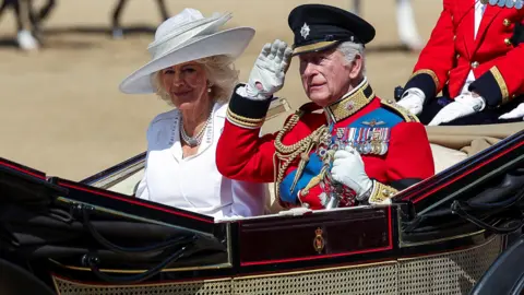King Charles, wearing a red military uniform adorned with medals, salutes with a white-gloved hand as he rides in a carriage alongside his wife Queen Camilla who is wearing a white dress and hat