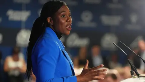 PA Media A woman with long hair wearing a bright blue jacket making a speech standing in front of a lectern