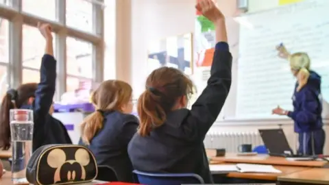 PA Media A back view of three girls in blue school jackets, two holding up their hands, looking towards a teacher writing on a whiteboard 