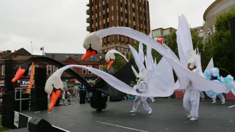 People dressed in enormous white and black swan costumes dance on a stage in a town centre