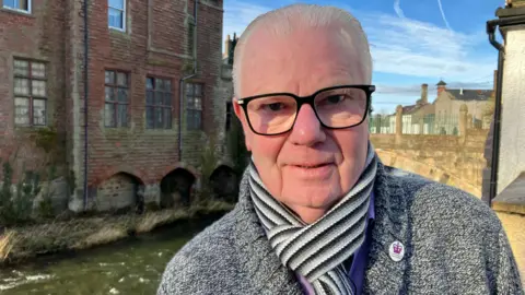 BBC/Ed Hanson Brian, who is clean shaven with short white and black glasses and is wearing a grey fleece and thick black and white scarf, stands beside a river.