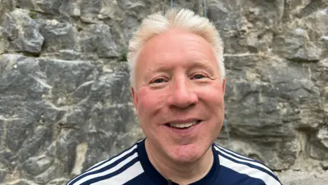 Older man with blond hair and a blue and white top in front of a rock face