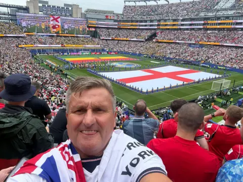 Ed Faulkner Ed Faulkner pictured ahead of kick-off at England's game against Ghana in the group stages. He is wearing a white England shirt with the flag draped over his shoulder.