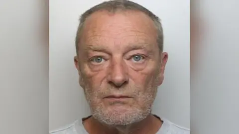 Paul Renwick looking at the camera in a police mugshot. He is an older man with short, graying hair and a beard or stubble. 