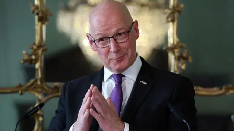 A bald man with glasses and wearing a black jacket, white shirt and purple tie has his hands pressed together as if in prayer