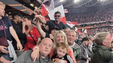A group of seven supporters at Old Trafford, all in the same row, smile for the camera. At the front is Katie, her husband and Ruairi.