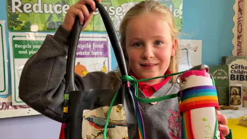 Ellen, a young pupil with blonde hair, holds up her black recycled bag with a patch of a blue bird on the front. Attached to the bag with green fabric is a reusable water bottle with a rainbow strap. It has 'reuse' written on the side in green felt tip marker.