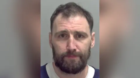 Norfolk Police Police issue picture of Adam Barnard. He is looking straight at the camera. He has short brown hair and is wearing a white t shirt and blue jumper. 