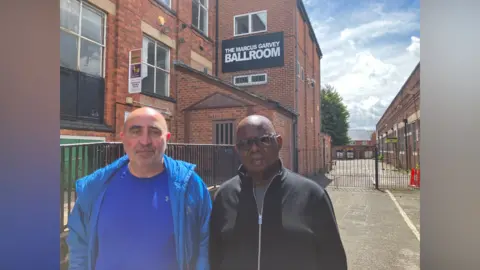 Two men stood outside the Marcus Garvey Ballroom building. Man on left in blue t shirt and jacket and man on right in black top and glasses.