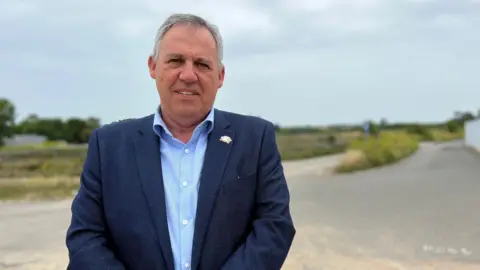 An image of Andy Jehan stood in front of a grassland and a path. He is wearing a blue suit shirt and a navy jacket. He has a pin badge on his suit jacket