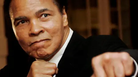 Muhammad Ali is seen throwing a mock punch in a close-up photo of his face and shoulders. He is wearing a white shirt and a black jacket