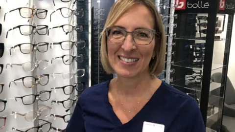 Rachael Smith is an optometrist. She has a blonde, bobbed hair and is looking towards the camera - smiling. She is inside the optometrists, standing beside a rack of glasses.