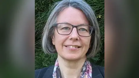 A woman with grey bobbed hair and black glasses wearing a flowery scarf and navy blazer. She is outdoors with greenery behind her and smiling.