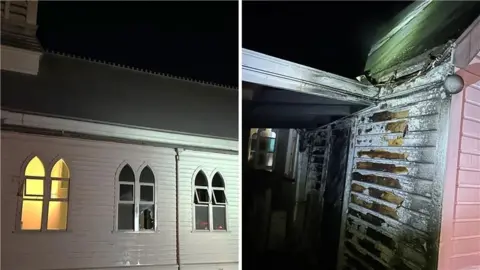 A white church illuminated by the glow of flames and a burnt wall are seen