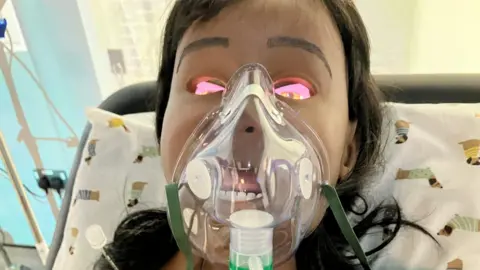 A manikin doll wearing an oxygen mask with brown hair leans back against a pillow