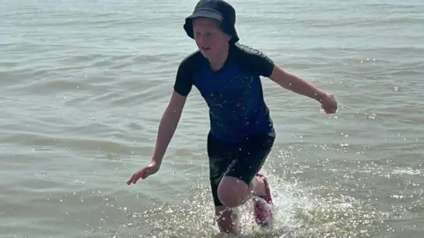 Family Theo runs through shallow sea water at a beach, wearing a dark top and hat as water splashes around his legs.