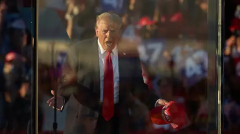 EPA Donald Trump with his hands apart, holding a Make America Great Again cap, on stage and behind bulletproof glass. He is wearing a red tie, white shirt and dark suit, in front of a crowd