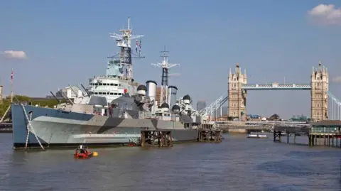 A World War Two era battleship moored on the River Thames in central London. Tower Bridge is in the background. 