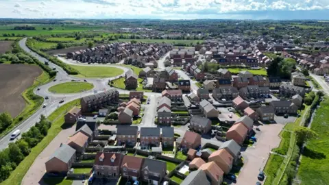 Drone footage of a new build housing estate. There is a large roundabout to the left and about 100 houses in the development. 