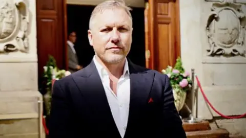 A man in his 50s, with short blonde hair and light stubble, looks into the camera with a serious expression. He is wearing a navy blue suit jacket over a white shirt, and is standing outside an ornate stone building with large wooden doors. 