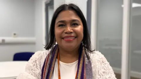 Domingas Gusmao is smiling at the camera. She has black hair which is just sitting on her shoulders. She is wearing a white top and lacy cardigan and a colourful scarf. She has pink lipstick on.