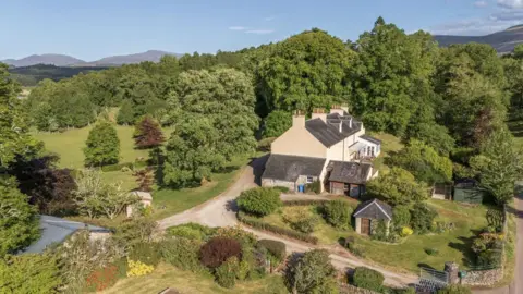 Galbraith The house is of period design with a slate roof and cream-coloured walls. It is set in a Highland landscape of rolling hills. Trees surround the property.