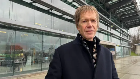 BBC Andy Gollifer in front of the National Glass Centre. He is wearing a navy blue coat, black jumper and a beige and black patterned scarf. He has straight brown hair.