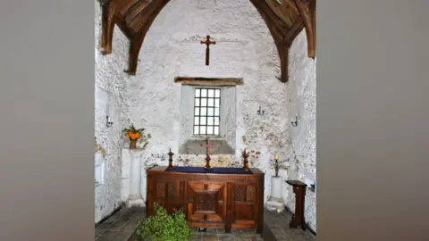 Cambridge Past, Present & Future Interior of Cambridge Leper Chapel, showing white washed walls, a wooden beamed roof above, a wooden altar below a window, with a cross hanging on the wall above it