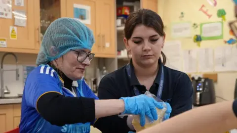 A woman wearing a blue hair net and blue gloves holds a plastic pot and is pouring its contents into an out of shot bowel. Another woman stands next to her with dark hair that has been tied back and is guiding her.