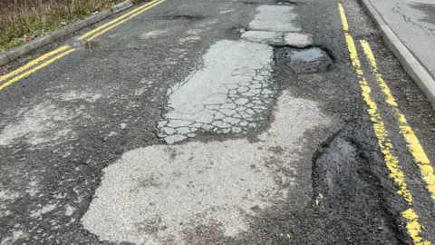 A narrow road with multiple potholes containing stones and pools of water. The surface is visibly poor and cracked, with multiple previous patches of the surface visible.
