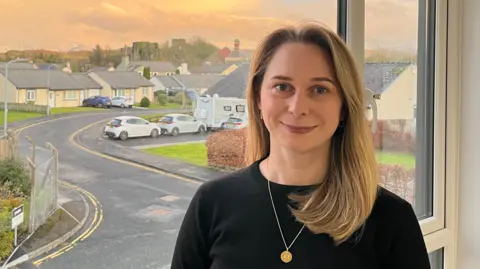 A head-and-shoulder image of Rosie Richardson. She has shoulder-length blonde hair and blue eyes. She is wearing a black jumper and a golden chain with a round pendant. She is standing in front of an office window at sunset. It looks out to a housing estate with Cockermouth Castle and Jennings Brewery visible in the skyline.