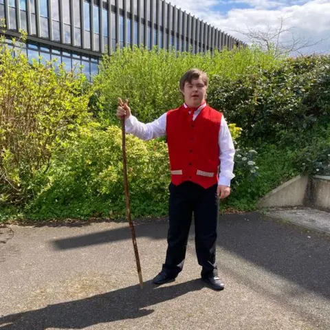 Sion holding a stick and wearing a red waist coat