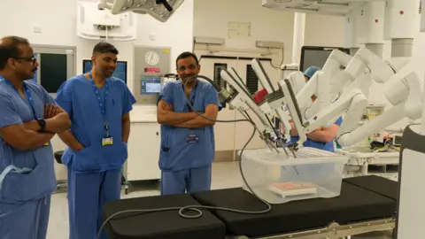 Three men in doctor's blue outfits look on at a surgical robot which is white and with a number of probes, they are all in what looks like an operating theatre