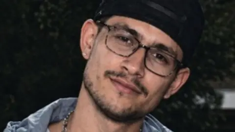 West Midlands Police Kristian Danielov is smiling, he is wearing a dark cap and glasses. A necklace can be seen around his neck and he is also wearing a light grey jacket. 