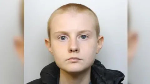 Alina Burns is a young woman with a shaved head. The image is a police mugshot. 