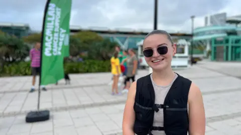 Meg is in the foreground wearing a black vest and grey top with 'PRO NIKE' on the waistband. Behind them is a green 'Macmillan Cancer Support' flag, several people in athletic clothing, and a building with glass windows under a cloudy sky.
