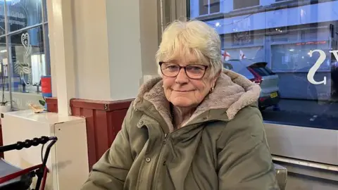 An elderly woman in a green coat. She is smiling. Behind her is car with England flags on it. 