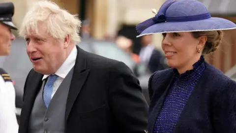 Boris and Carrie Johnson appear next to each other. Boris wears a black suit, grey waistcoat and blue tie, while Carrie wears a dark blue dress and lighter blue hat.