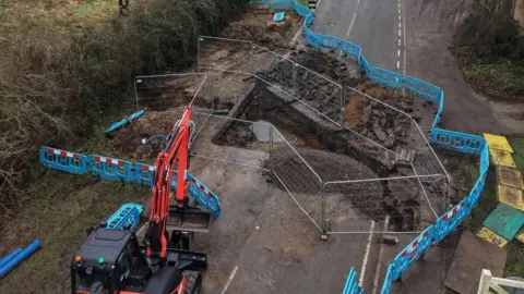Part of a road is blocked off with blue and silver barriers, with a large hole in the tarmac and a red and black digger parked nearby.