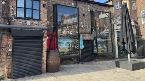 Outside of a bar. The photo is taken in the daytime and the sky in the background is blue. The building is a medium size brick building with black framed windows. There are several large pub-garden style umbrellas unopened in shot. One window has 'Trader's' printed in white on the glass.