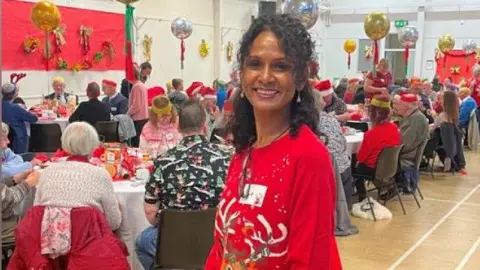 A woman with dark brown hair is wearing a red Christmas jumper with a brown reindeer on the front. She is smiling. Behind her, in a sports hall, there are long tables with dozens of people sitting at them, also wearing Christmas jumpers. There are gold and silver balloons hanging up