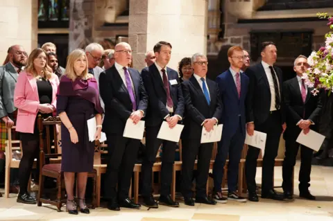 MSPs and guests stand holding service programmes during the Royal Kirking of Parliament at St Giles' Cathedral in Edinburgh. The group, dressed in formal attire, faces the front of the cathedral during the ceremonial church service beneath the building’s stone arches and warm lighting.