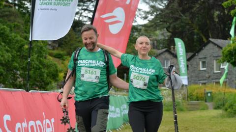 Jeremy Langmead and Sarah Edmundson, both in green Macmillan charity shirts, crossing the hike finish line.