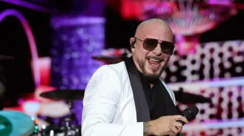 Rapper, Pitbull, performing at a concert. He is wearing a white suit jacket, a black shirt underneath and black sunglasses. He has a microphone in his right hand, and a black earpiece in his right ear. Behind him is the concert set with a digital screen which has graphics on it. 