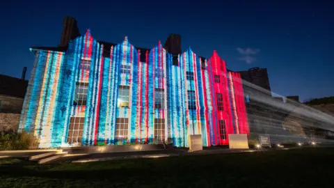 The graphic showing the stripes, starting blue on the left hand side and changing to deep red on the right hand side, being projected onto the stone wall of a castle at night